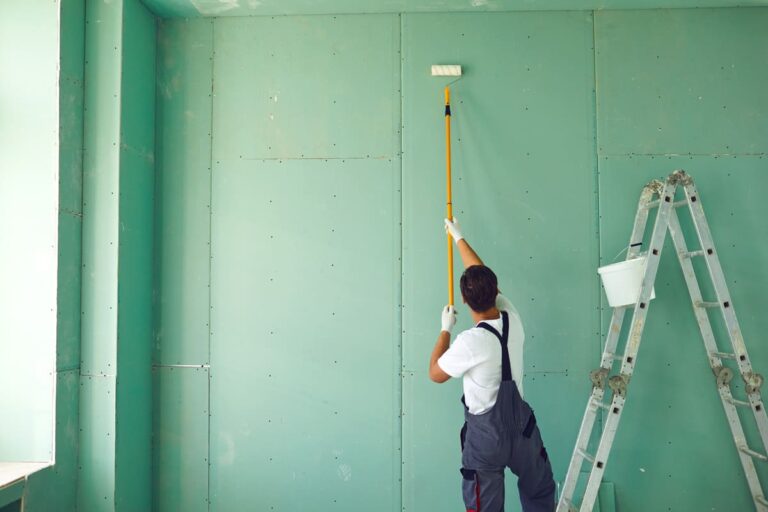 L'image montre un ouvrier d'une entreprise de peinture professionnelle, en train de peindre un mur intérieur vert clair à l'aide d'un rouleau monté sur un manche télescopique. L'homme, vu de dos, porte une combinaison de travail grise et un t-shirt blanc, et semble concentré sur sa tâche. À sa droite, une échelle en métal est posée contre le mur, avec un seau blanc suspendu contenant probablement des outils ou de la peinture. La pièce en cours de rénovation est recouverte de plaques de plâtre, soulignant l'état de progression des travaux. Cette scène illustre un processus de rénovation intérieure, mettant en avant le travail de peinture et de préparation des surfaces.